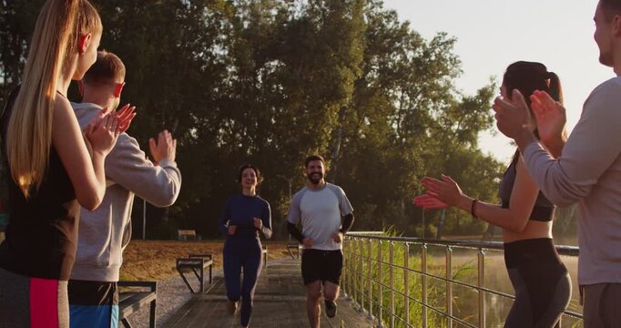Group of cheerful people stands in summer park, applauding and giving high-fives to two joggers just finished run at finish line, congratulating, supporting, exuding motivation and success