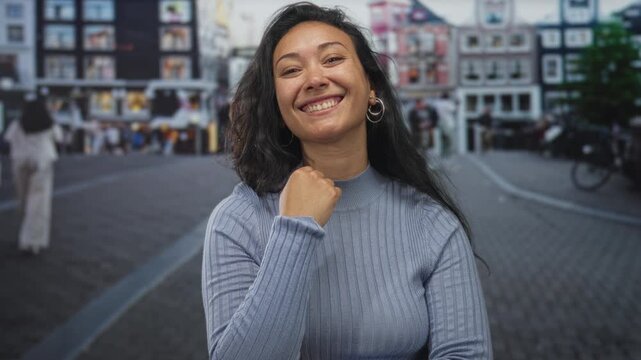 Young hispanic woman smiling and holding fist under chin on a crowded cobblestone street with blurred buildings and bicycles; confidence.