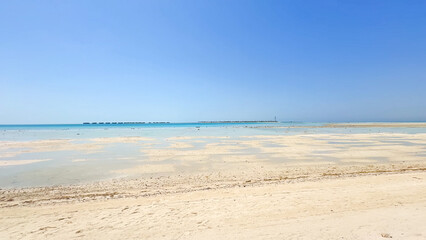 Vast expanse of beach and shallow, receding water under a clear blue sky, with distant water villas on the horizon