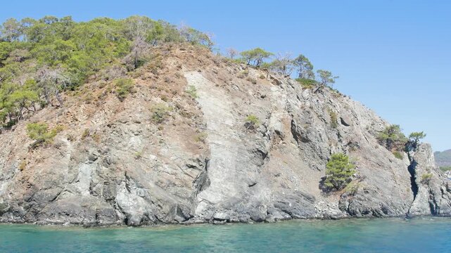 A stunning cinematic shot of a rugged, rocky coastline where steep cliffs covered in lush Mediterranean maquis (shrubland) meet the crystal-clear turquoise waters.