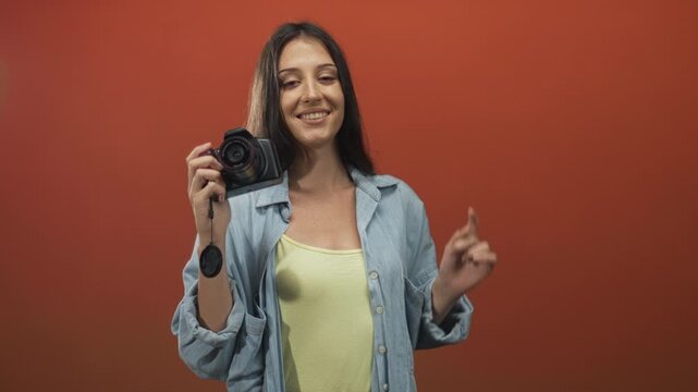 Woman holds camera up while smiling and pointing with right hand in red studio wearing denim shirt and yellow tank top; joy creativity.