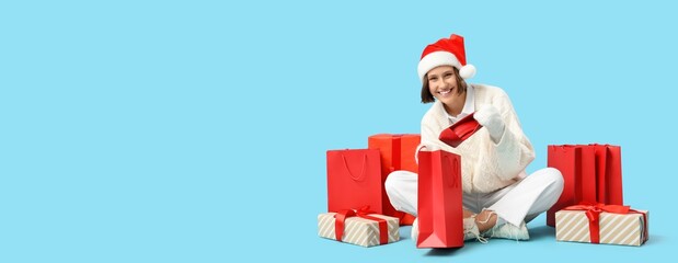 Happy woman in Santa hat with shopping bags and gifts on blue background with space for text. Christmas sale