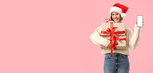 Young woman in Santa hat holding gift boxes and mobile phone on pink background with space for...