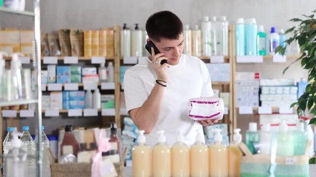 Guy is holding a package of pads in his hands and calls his wife on the phone to clarify which ones to buy. Young man buying pads for his wife at the pharmacy. High quality 4k footage