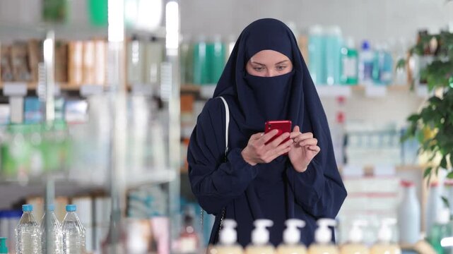 Young Muslim woman in traditional islamic navy blue dress and niqab covering face except eyes, shopping for medications or care products in drugstore, using phone to check list of needed items