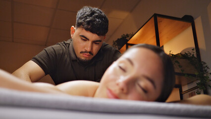 A man in a black shirt massages a woman's back in a serene, warmly lit room, suggesting a professional spa environment. The woman appears calm and relaxed, emphasizing the therapeutic.
