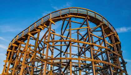 Low-angle view of a vibrant orange wooden roller coaster track against a clear blue sky with wispy clouds.