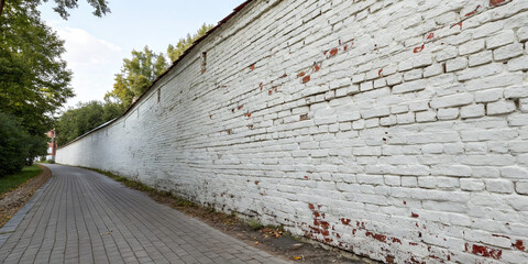 A long, white brick wall stretches alongside a paved path in a park. Lush trees frame the scene, providing a calm, natural atmosphere under the blue sky