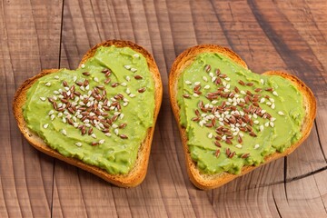 Two heart-shaped toasted bread slices spread with avocado paste and sprinkled with flax and sesame seeds on a wooden background
