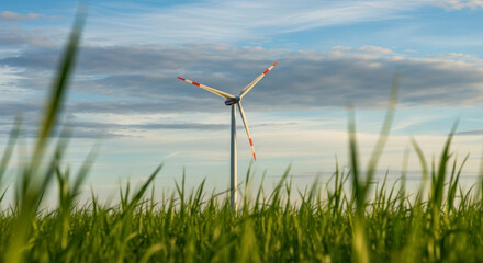 Wind turbine with red-tipped blades standing tall in a vibrant green field of grass under a partly cloudy blue sky in the evening light