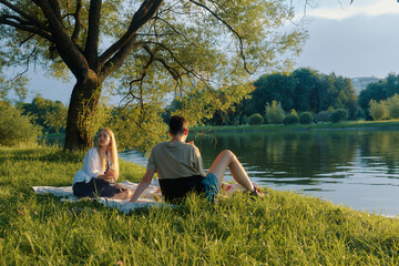 Man and young woman enjoy a picnic by a tranquil pond under the shade of a tree on a sunny day