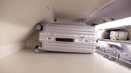 Passenger placing silver trolley suitcase into overhead luggage compartment inside economy class airplane, then closing bin door. Camera placed inside compartment