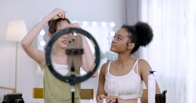 asian teenage girl adjusting hair rollers while african american teenage girl looking and smiling during beauty tutorial shoot at home studio fun influencer friendship lifestyle cosmetic content