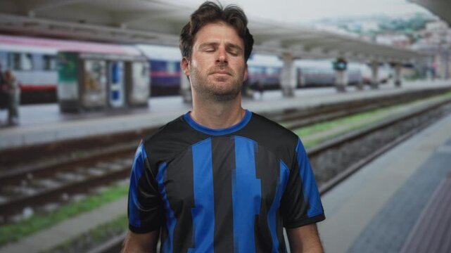 Man in blue soccer jersey smiling at camera on train platform inside a station building near railway tracks; contentment travel.