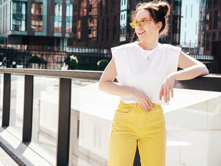 Fototapeta premium Young beautiful smiling hipster woman in trendy summer yellow jeans clothes. Carefree teen model posing in the street at sunset. Positive female outdoors. Cheerful and happy. In sunglasses