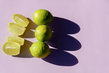 Fresh green limes and slices arranged on a vibrant purple surface, creating strong contrasting shadows in bright sunlight