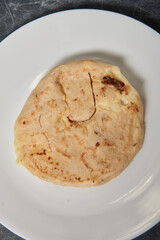 A close-up photo of a golden, cheese-filled pupusa on a white plate against a black-grey marble background.