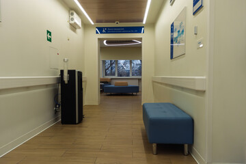 Clean hospital corridor featuring blue seating benches, handrails, informative signs, and a water purifier. Leads to a waiting area with large windows and additional seating