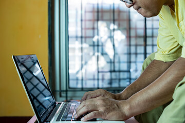 A focused middle-aged man in a yellow shirt and glasses works on a laptop at a desk near a window, concentrating on typing. The setting features bright yellow wall with natural light.