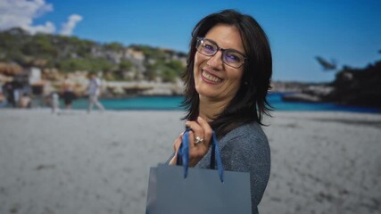 Smiling woman holding shopping bag handle with raised hand at sunny beach coastline; happiness relaxation. - Powered by Adobe