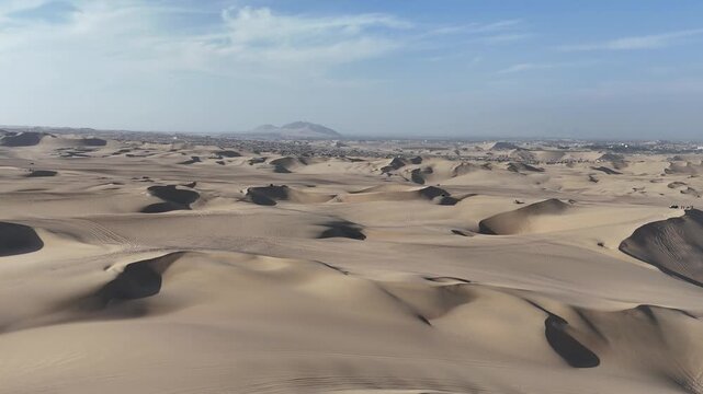 Aerial moves forward over Huacachina's light orange sand dunes under a blue sky, capturing multiple dune buggies driving fast across the desert landscape