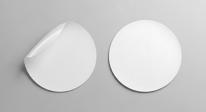 Two blank white round stickers on a gray background, one with a peeled-up edge, suggesting adhesive
