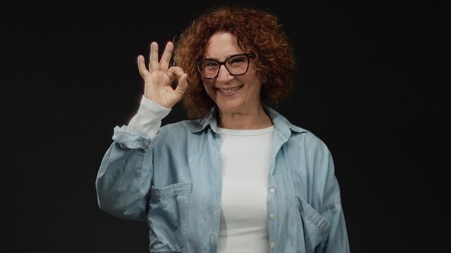 Middle-aged woman with curly hair wearing glasses and casual attire shows an ok gesture against a black background, exuding confidence and positivity.