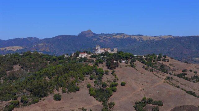 Aerial drone photo of Hearst Castle located on a hill near San Simeon California surrounded by dry hills and greenery under clear blue sky showcasing historic architecture and coastal landscape