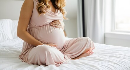 A pregnant woman sits on a bed, cradling her baby bump with her hands, wearing a soft pink maternity dress