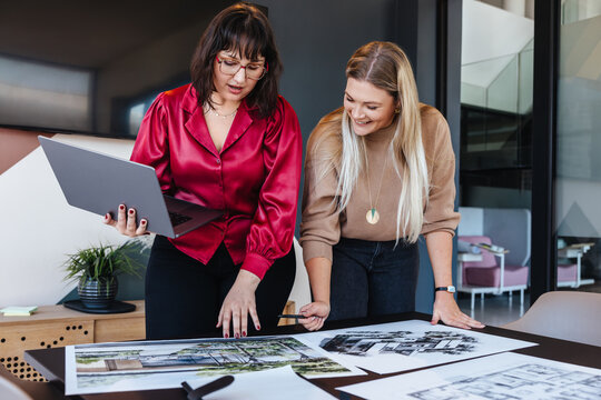 Two women collaborating on architectural designs at a modern workplace using a laptop - Powered by Adobe