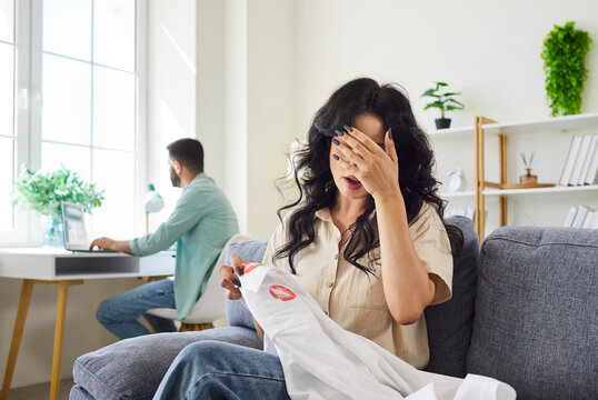 Shock and stress of wife holding husbands white shirt with red lipstick and kiss stains. Hands of woman cover eyes to avoid seeing evidence of infidelity, adultery and betrayal of unfaithful man