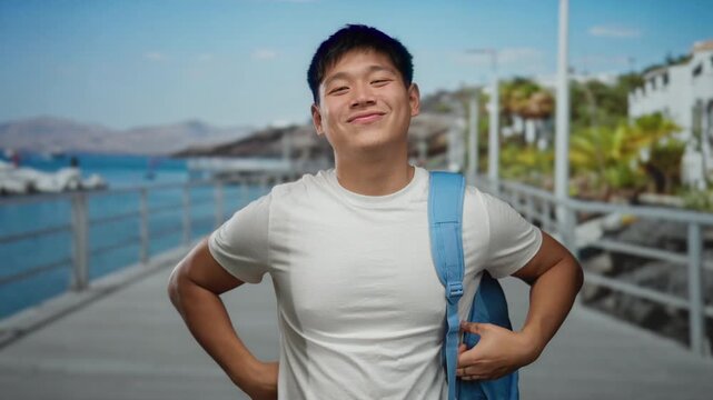 Young man smiling and posing with peace signs near scenic seaside promenade, wearing casual white shirt and blue backpack, with blurred beach and ocean background under clear sky.