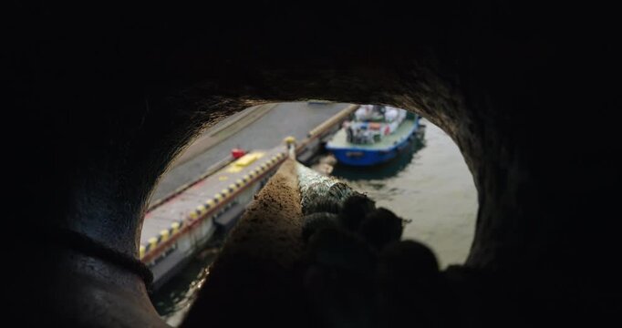 View through circular opening of ship hawsehole frames scene of dock and small vessel. Weathered mooring rope leads eye to bright daylight