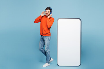 Full length young man wear orange sweatshirt casual clothes big huge blank screen mobile cell phone smartphone with copy space mockup area listen music in headphones isolated on plain blue background.