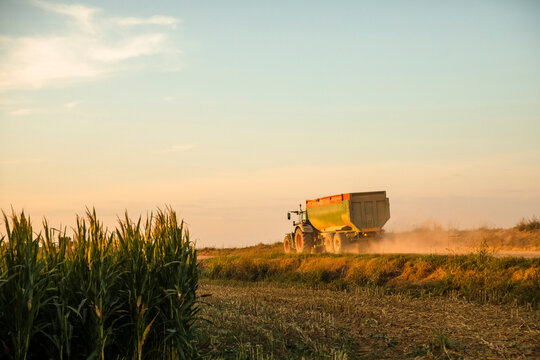 Tractor pulling trailer on rural farm road at sunset with dust rising