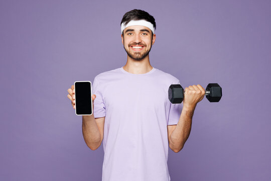 Young happy strong fitness trainer sporty man wear t-shirt train in home gym hold dumbbell use blank screen area mobile cell phone isolated on plain purple background. Workout sport fit abs concept.