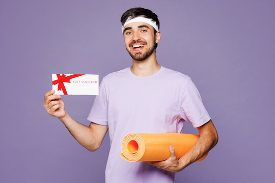 Young happy strong fitness trainer sporty man sportsman he wear t-shirt train in gym hold yoga mat gift coupon voucher card for store isolated on plain purple background Workout sport fit abs concept