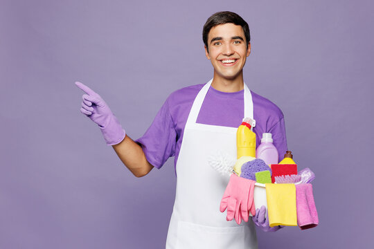 Young man in violet t-shirt hold basin with detergent bottles while do housework tidy up point index finger aside on area isolated on plain pastel light purple background studio Housekeeping concept