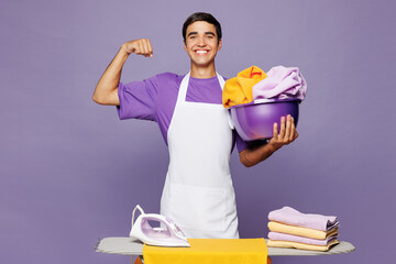 Young smiling happy man he wear violet t-shirt iron clothes on ironing board while doing housework tidy up show muscles biceps isolated on plain pastel light purple background. Housekeeping concept.