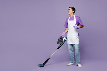 Full body side view young man wear violet t-shirt cleaning room using modern vacuum cleaner while...