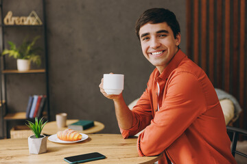 Young man he wear orange casual clothes hold in hand cup drink tea sits alone at table in coffee shop cafe relax rest in restaurant during free time indoors. Freelance mobile office business concept.