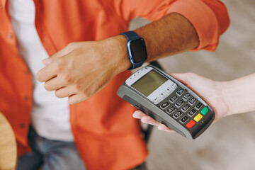 Close up cropped young man holding bank payment terminal process acquire smart watch sits at table in coffee shop cafe relax rest in restaurant in free time indoors. Freelance mobile office concept.