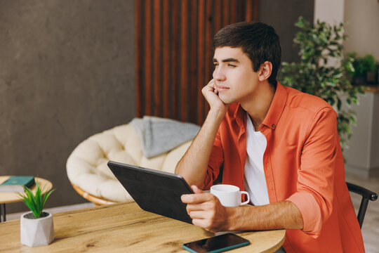 Young IT man he wears orange casual clothes digital tablet pc computer sitting at table in coffee shop cafe relax rest in restaurant during free time indoors. Freelance mobile office business concept. - Powered by Adobe
