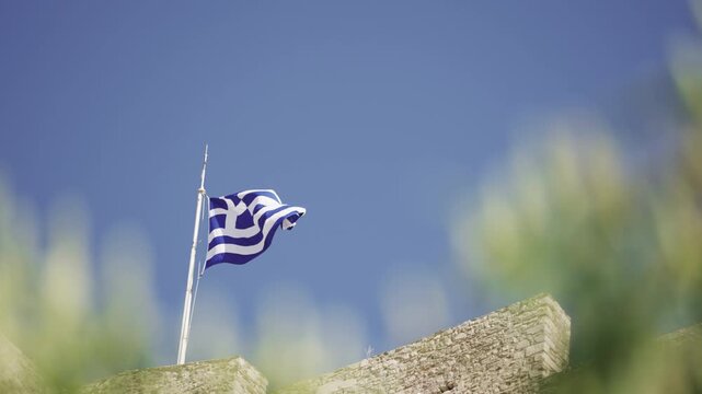 Greek flag waving on pole at ancient Acropolis in Athens Greece on column remains against blue sky background, spring sunny day, slow motion