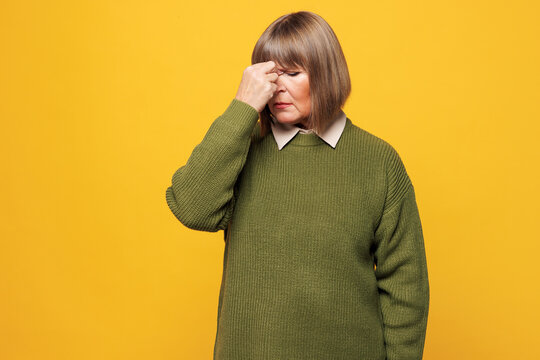 Elderly sad sick ill tired weary blonde woman she wear green sweater casual clothes keep eyes closed rub put hand on nose isolated on plain yellow orange background studio portrait. Lifestyle concept. - Powered by Adobe