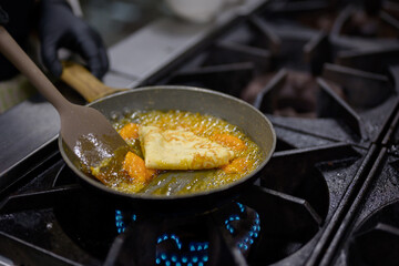 Chef cooking orange butter sauce for Crepes Suzette on gas stove, dessert caramelization process in professional kitchen environment.