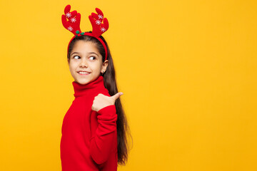 Merry little girl wear red clothes fun decorative deer horns on head posing point thumb finger aside on area isolated on plain yellow background. Happy New Year Christmas celebration holiday concept.