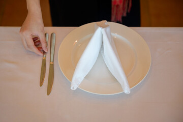 A close-up photo of a hand arranging silver knives beside a white plate with a folded napkin on a white tablecloth, shot in warm ambient light with shallow depth of field.