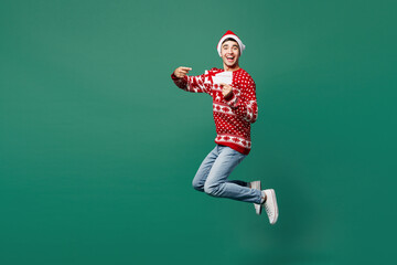 Full body merry young man wear red Christmas sweater Santa Claus hat posing jump high hold store...
