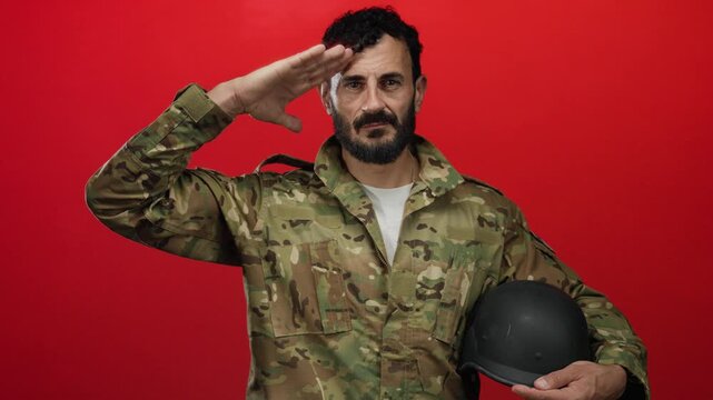 Military man in camouflage uniform gives a salute against a vibrant red background while holding a helmet in his other hand, showcasing dedication and service.